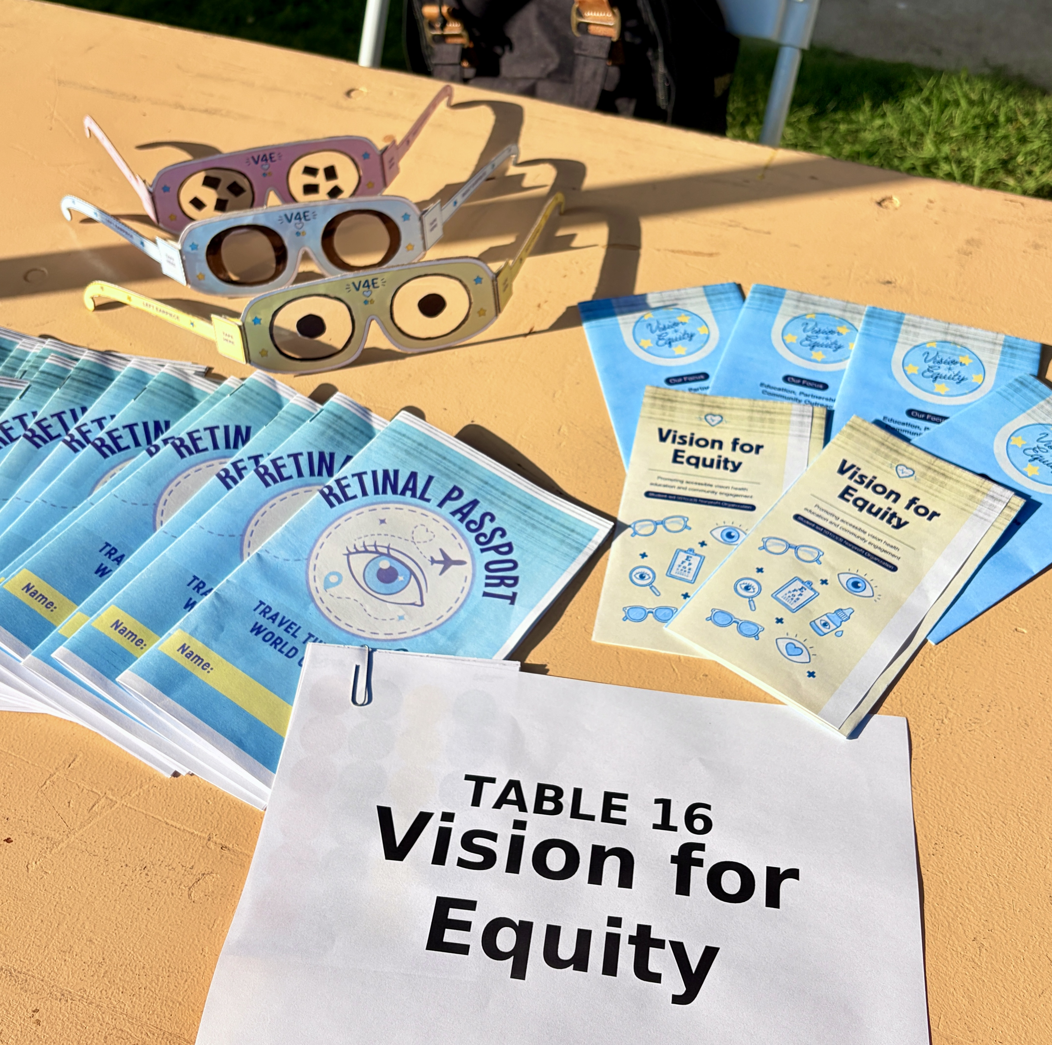 Vision for Equity outreach table with paper glasses, Retinal Passport booklets, educational brochures, and a Vision for Equity sign